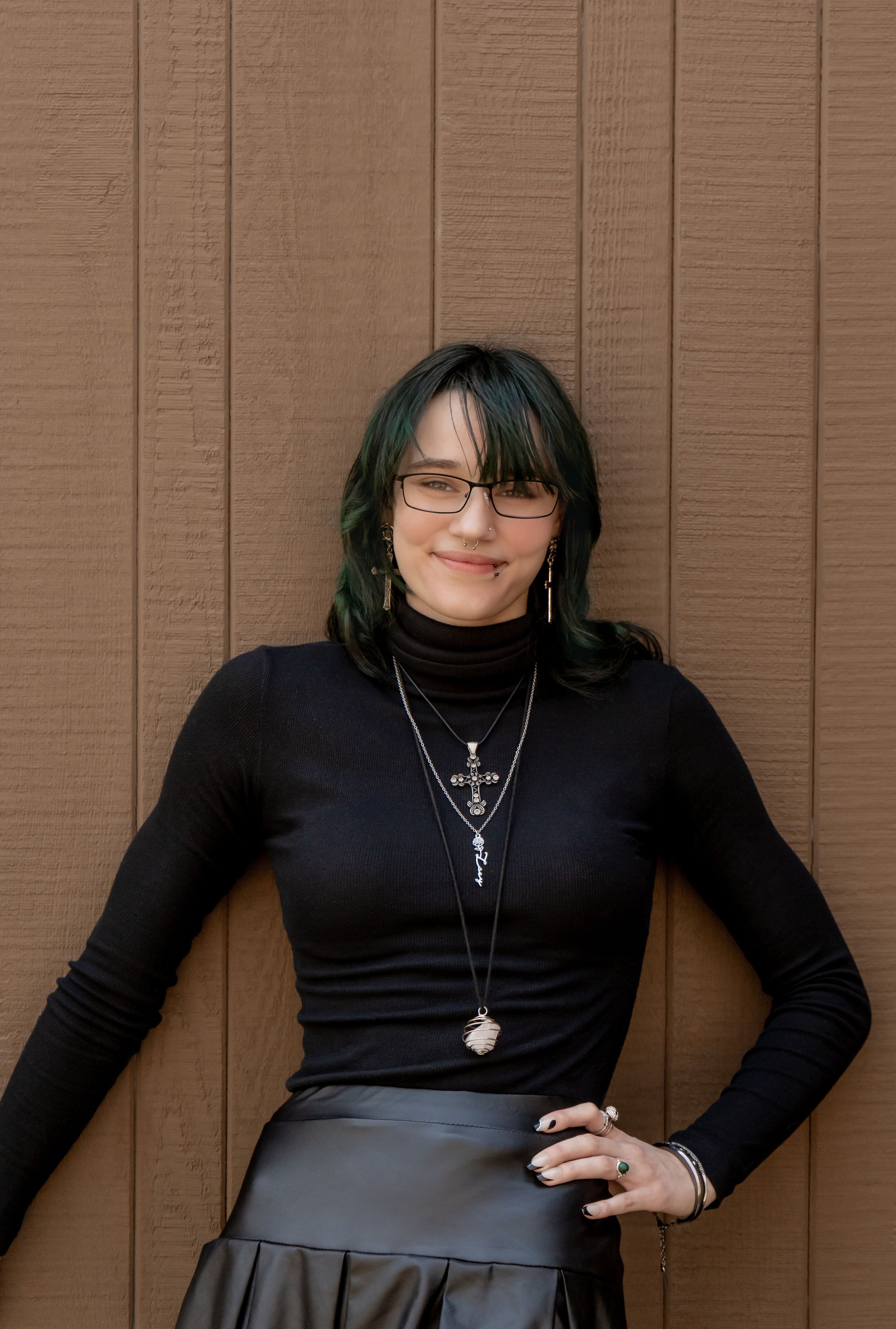 Woman with green-tipped hair in black turtleneck and skirt leaning against brown wall.