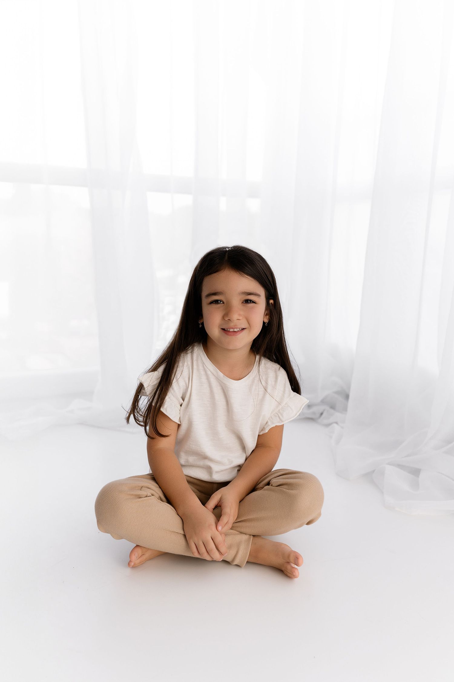 Young girl in cream shirt and khaki pants sitting cross-legged on white studio floor.