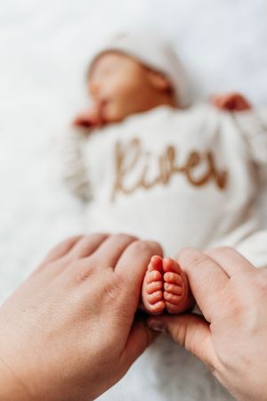 Parent hands gently holding tiny newborn baby feet with sleeping infant named River in background.