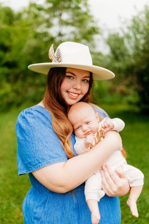 Smiling mother in white hat and blue dress cuddling her infant baby in a green garden.