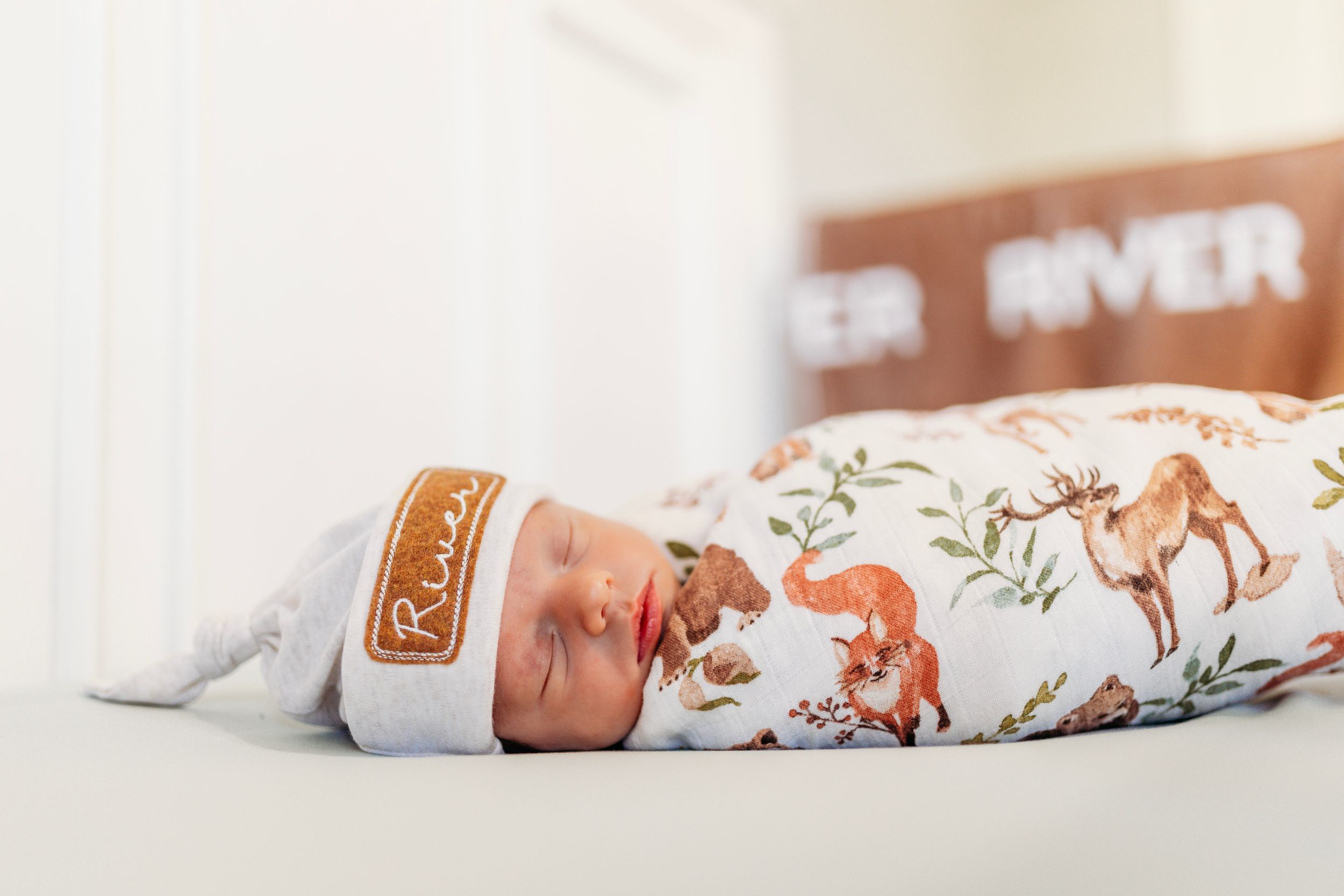 Newborn baby named River swaddled in woodland animal print blanket wearing personalized name hat.