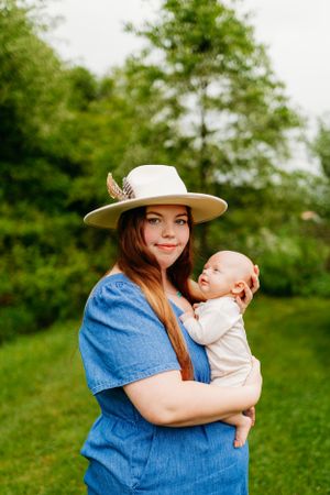 Woman in white hat and blue dress holding a baby outdoors in a lush green garden.