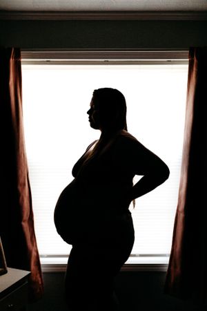 Dramatic silhouette of pregnant woman with baby bump standing in front of bright window indoors.