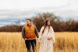 Expecting couple holding hands in golden autumn field under cloudy sky, woman cradling baby bump.