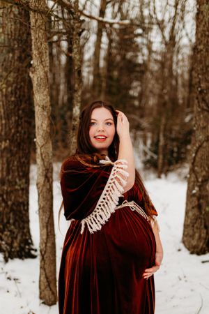 Smiling pregnant woman in burgundy velvet dress and fringe shawl posing in snowy winter forest.