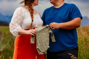 Couple holding small green floral onesie in golden field, announcing pregnancy outdoors with mountains behind.