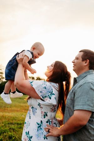 Happy family at golden sunset, mother lifts smiling baby while father stands beside them in a field.