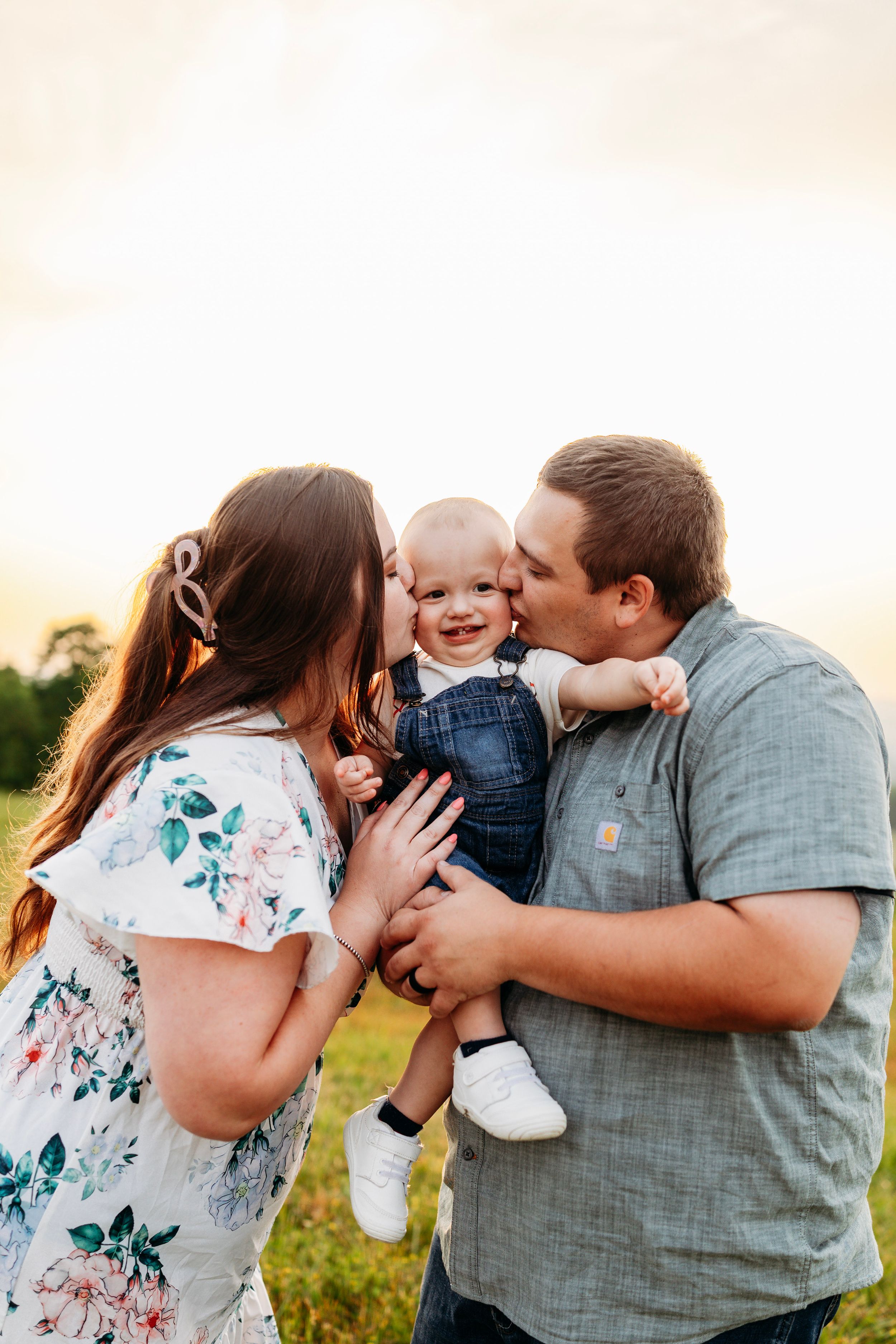 Parents simultaneously kiss their smiling baby on both cheeks during a warm golden sunset field session.