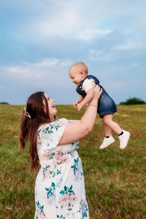Joyful mother in floral dress lifts laughing baby overhead in a sunny green meadow under blue sky.