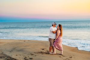 Couple embracing on a sandy beach at sunset with colorful sky over the ocean.