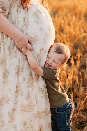 Happy toddler boy hugging pregnant mothers belly in golden sunset wheat field.