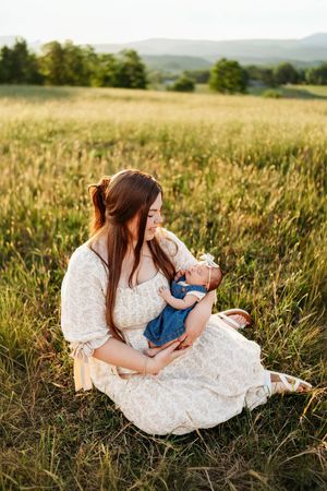 Mother in floral dress sitting in sunny field cradling smiling newborn baby girl.
