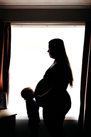 Silhouette of pregnant woman standing by bright window with young child touching her baby bump.