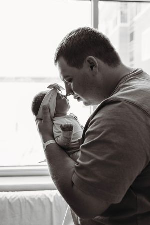 Black and white photo of father tenderly holding newborn baby girl by hospital window.