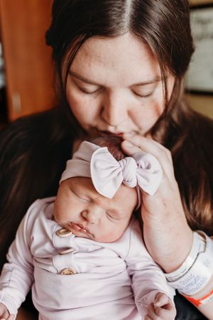 New mother kissing sleeping newborn baby girl wearing white bow headband in hospital.