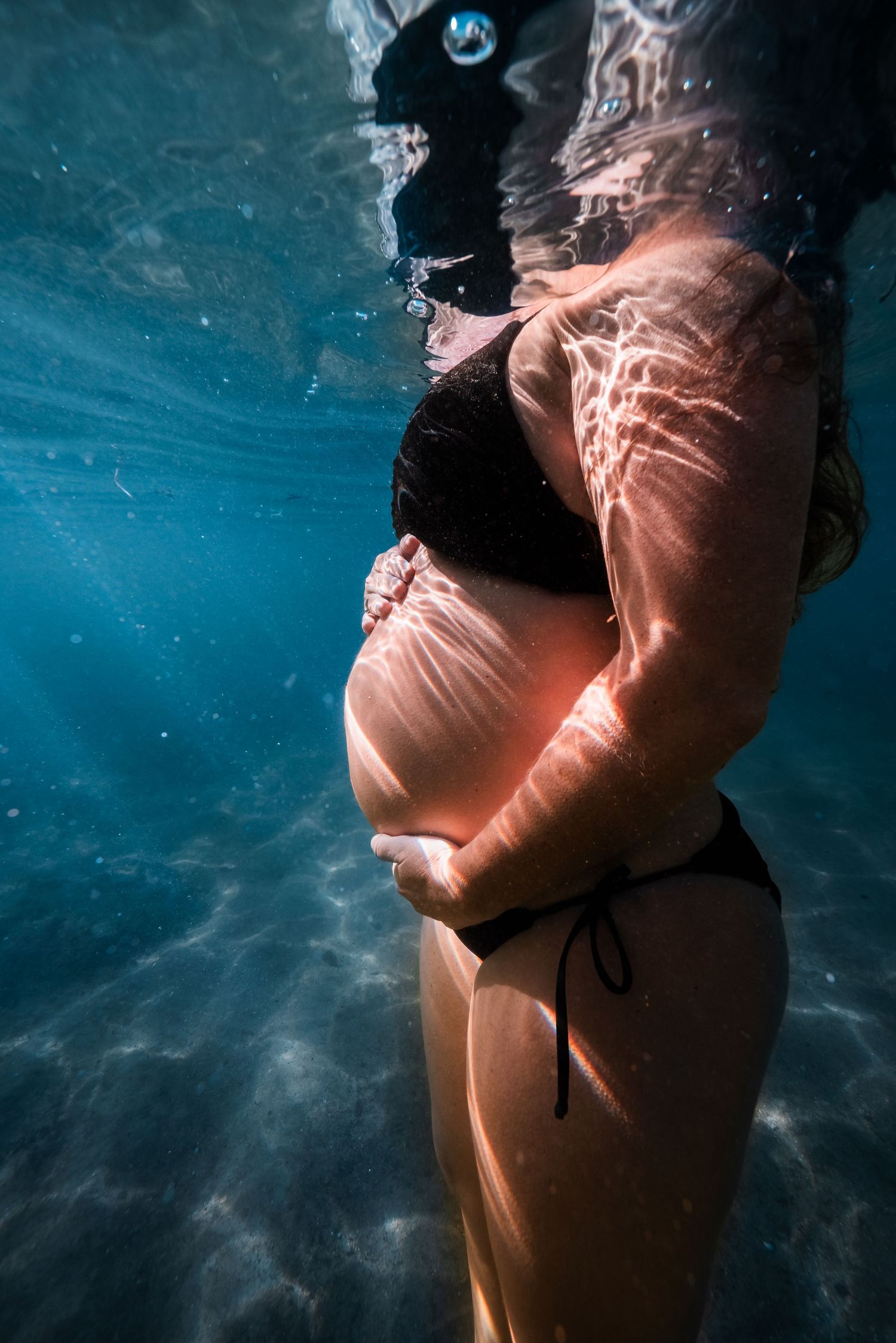 Pregnant woman in black bikini cradling her baby bump underwater, with light rays filtering through clear blue ocean water.
