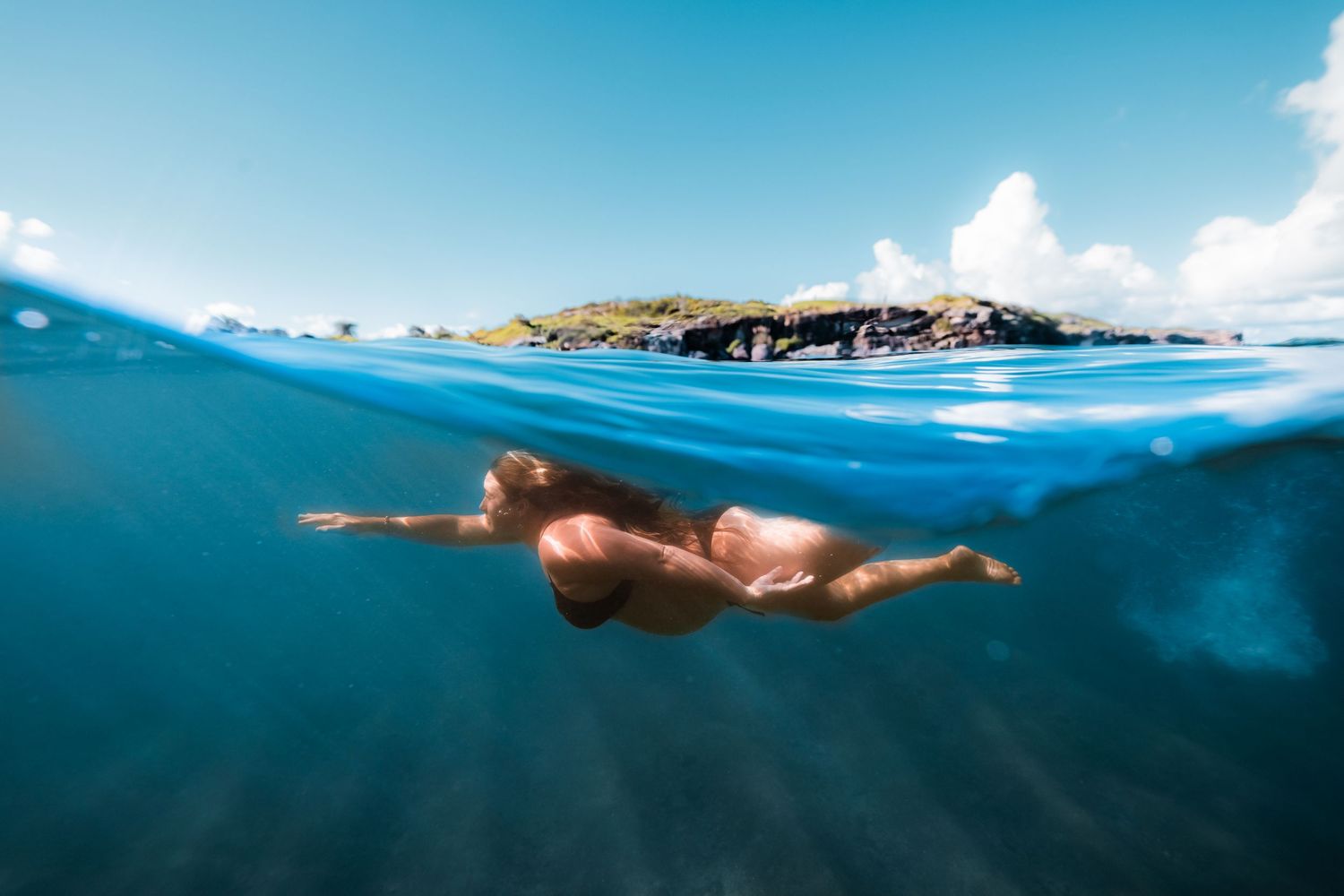 Woman swimming underwater in crystal clear tropical blue ocean near a lush green island shore.