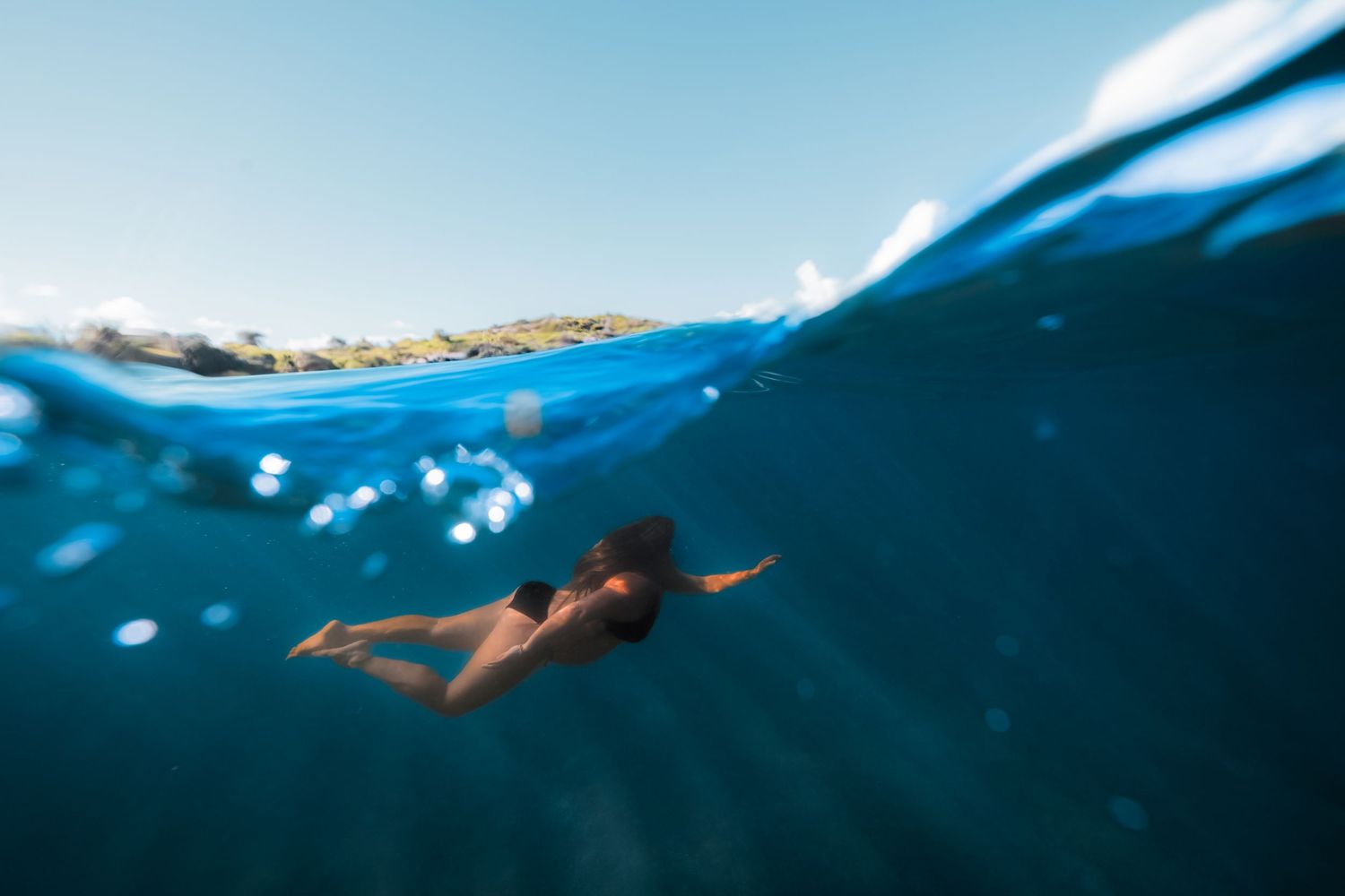 Underwater view of a woman snorkeling in clear blue tropical ocean water near a rocky shoreline.