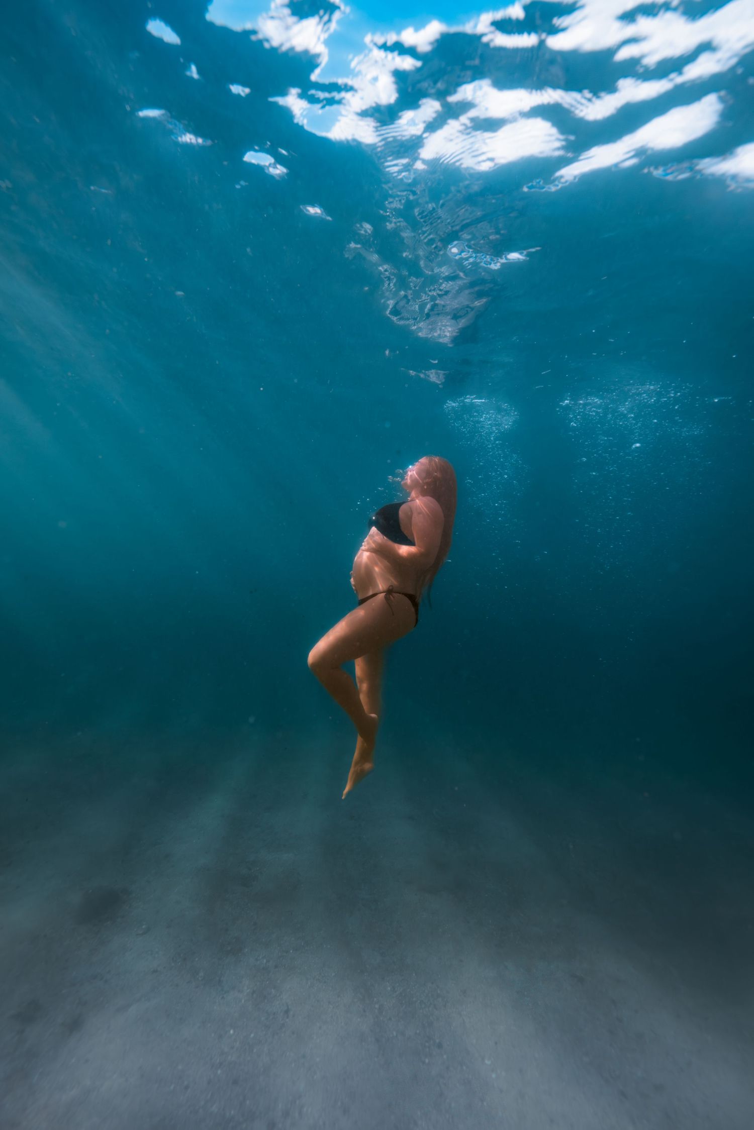 Underwater photo of a woman in a black bikini swimming in clear blue ocean water above a sandy seabed.