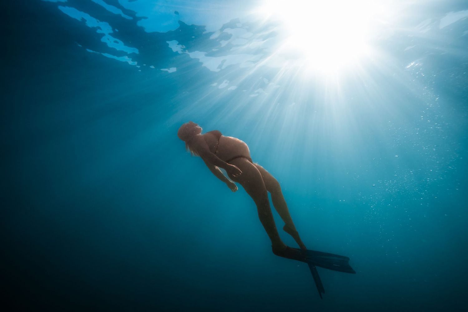 A silhouetted swimmer glides beneath sunlit turquoise waters with sunbeams streaming through the surface.