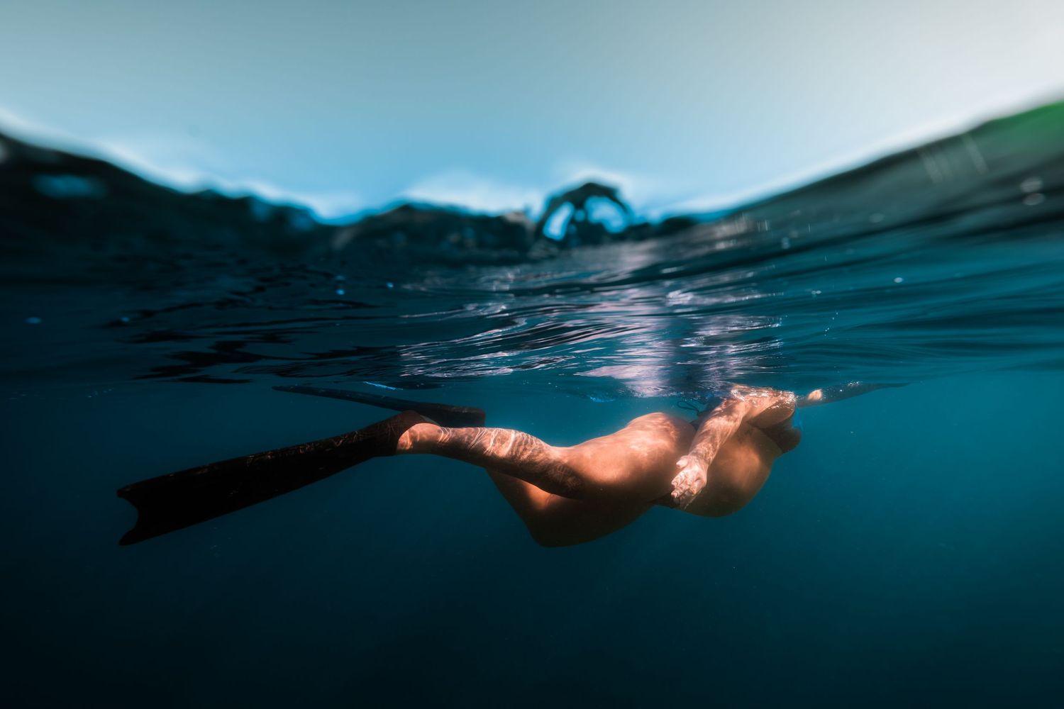 Underwater view of someone free diving in dark blue ocean water under sunlit surface.
