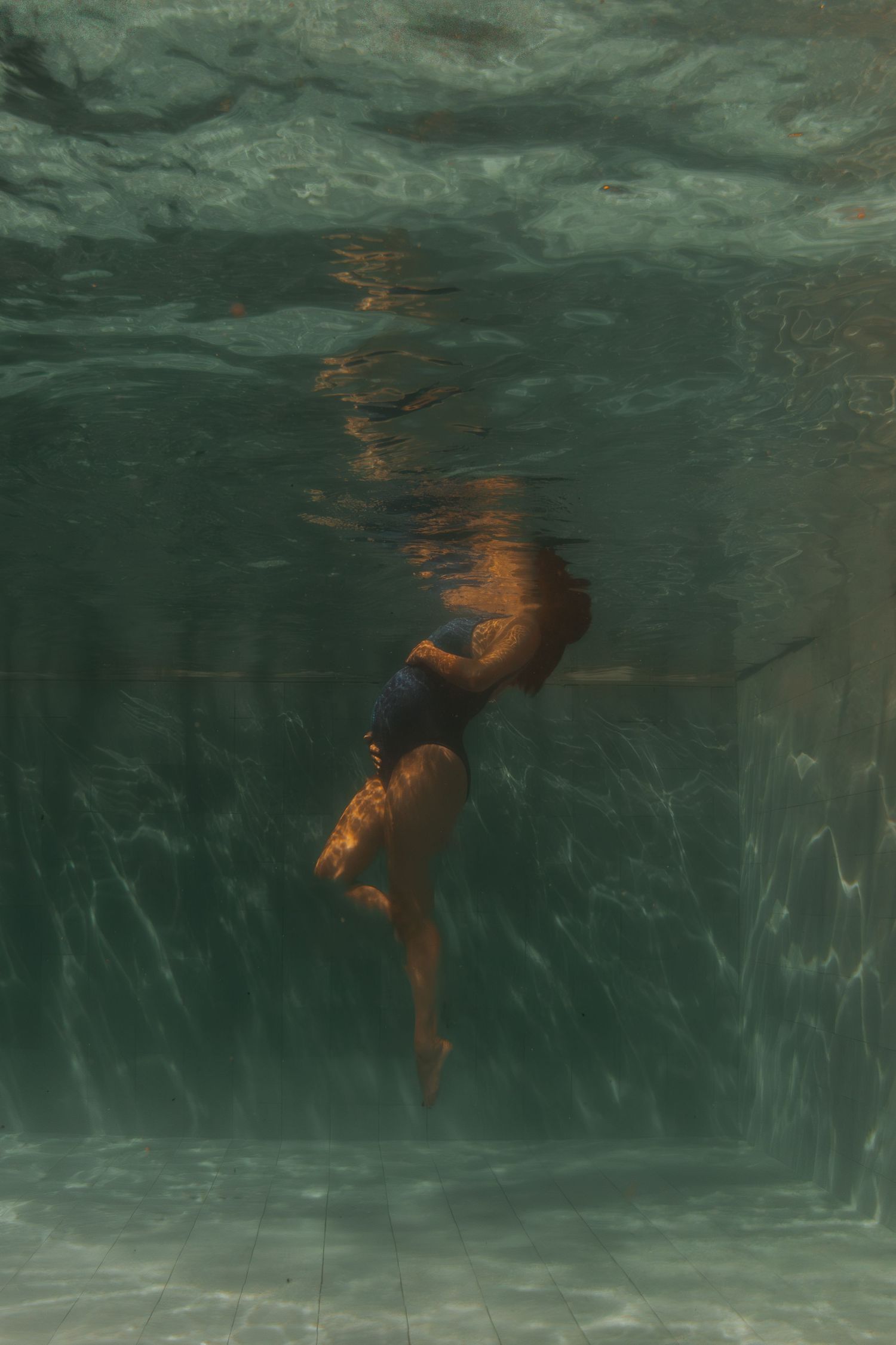 Figure in a black swimsuit floating underwater in murky green water, creating a moody atmospheric scene.