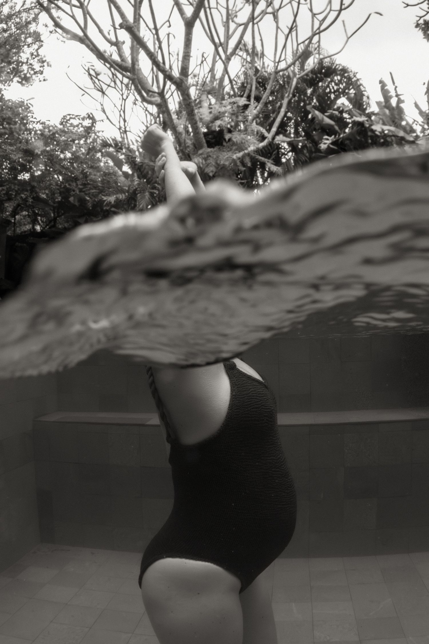 Black and white underwater photograph of a figure in a one-piece swimsuit with trees visible above the water line.