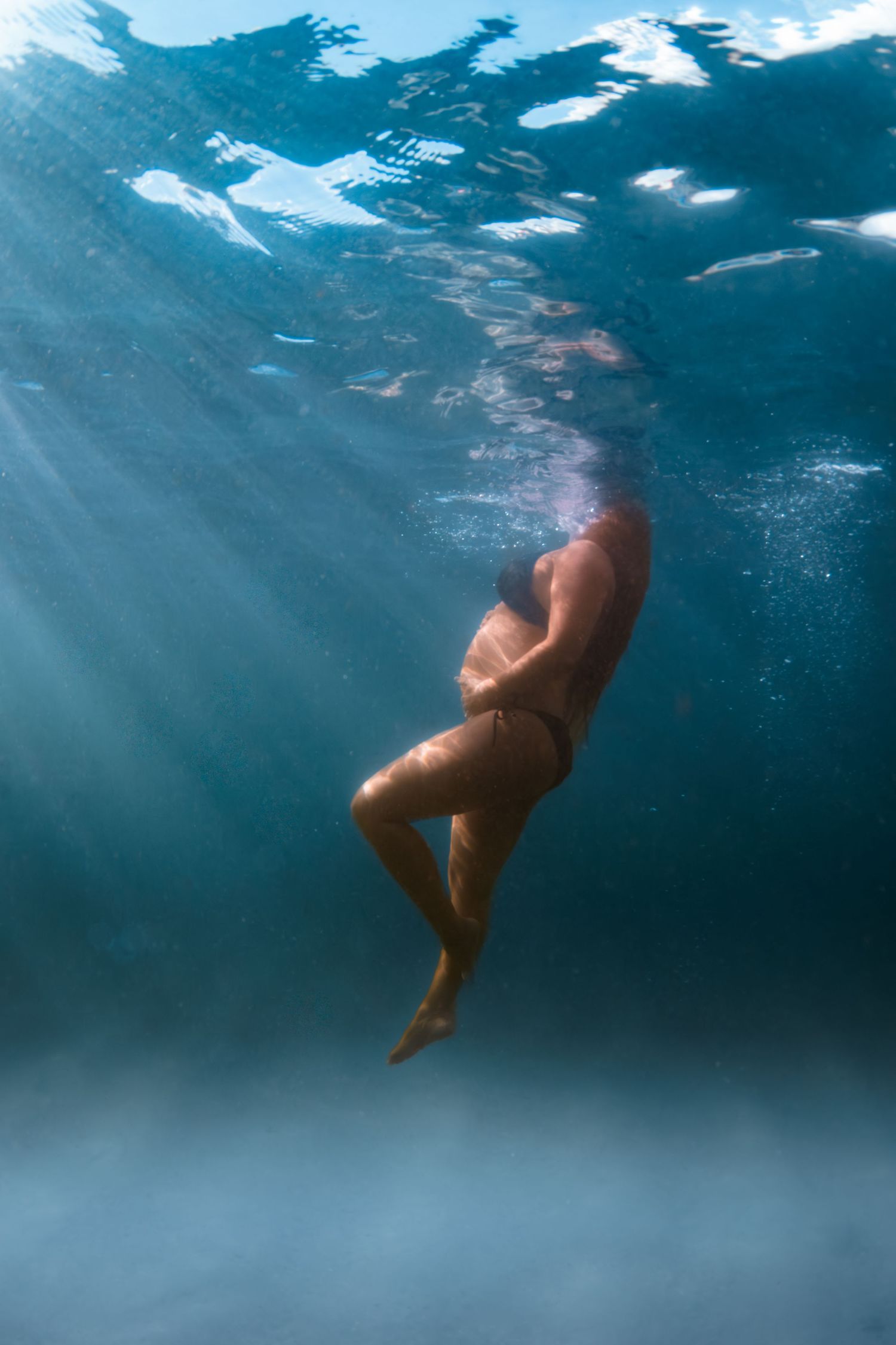 Underwater view of a person in a bikini swimming near the surface with sunlight rays filtering through clear blue water.