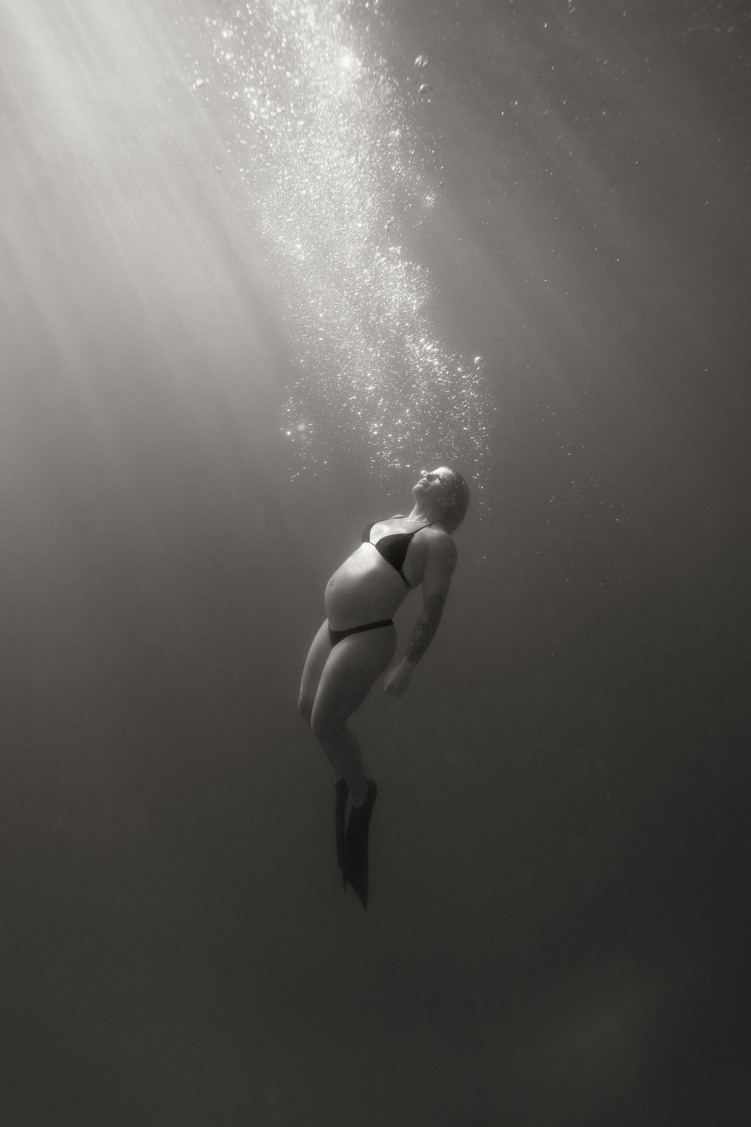 Black and white underwater photo of a swimmer floating in beams of light with rising bubbles in dark water.