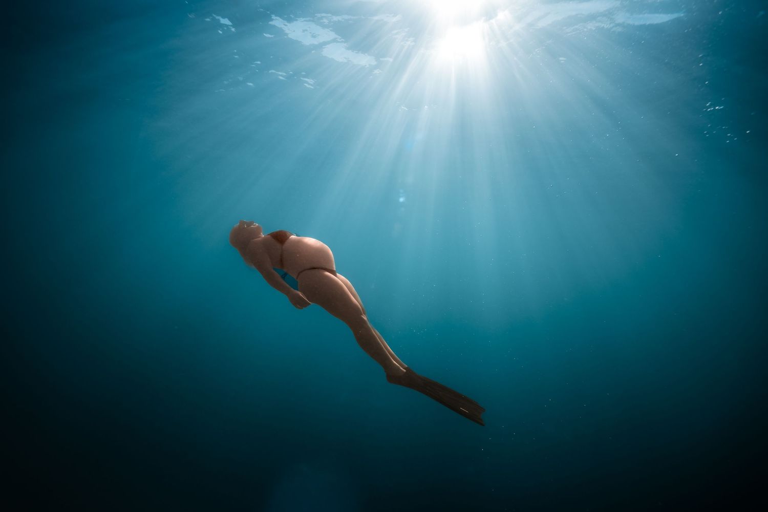 A silhouetted swimmer glides through deep blue underwater rays of sunlight filtering down from above.