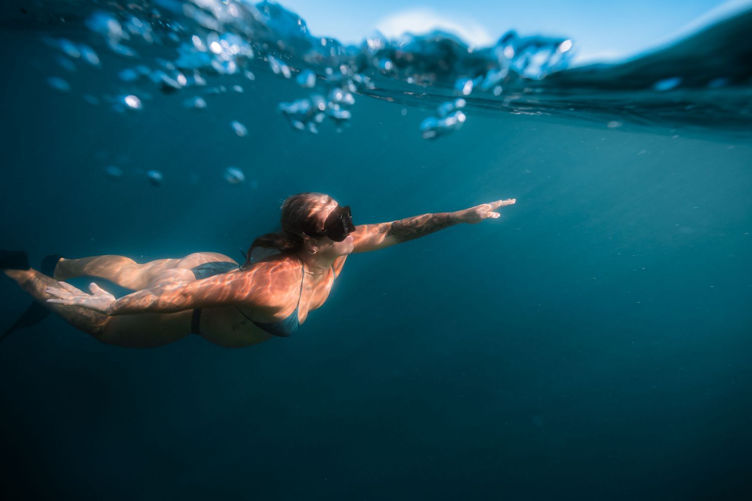 Swimmer gliding underwater with outstretched arms in turquoise blue waters beneath a rippling surface.