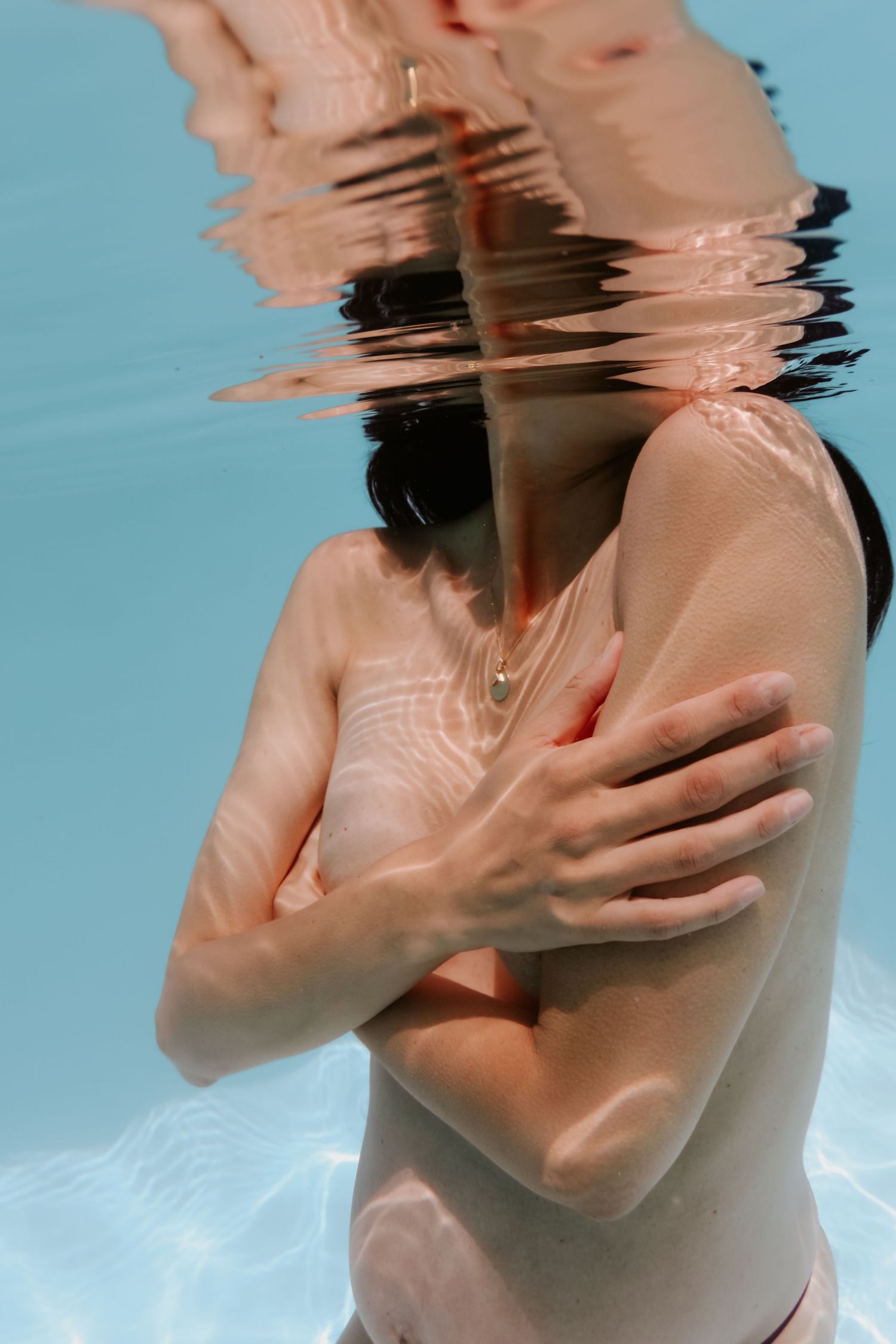 Person submerged underwater in a pool, arms crossed over chest, with light refracting through the water surface.