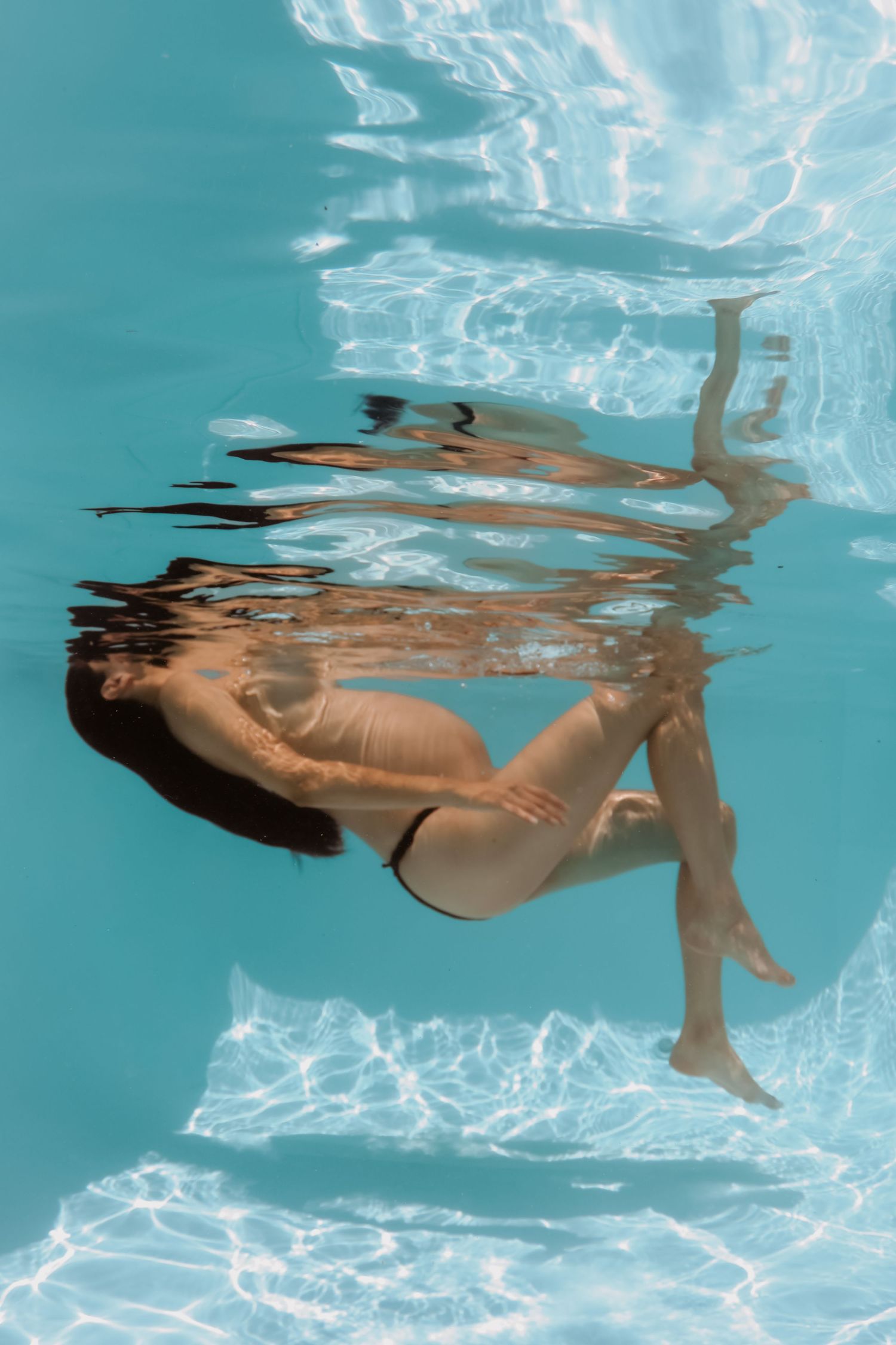 Pregnant woman in black bikini posing elegantly underwater in a bright blue swimming pool.
