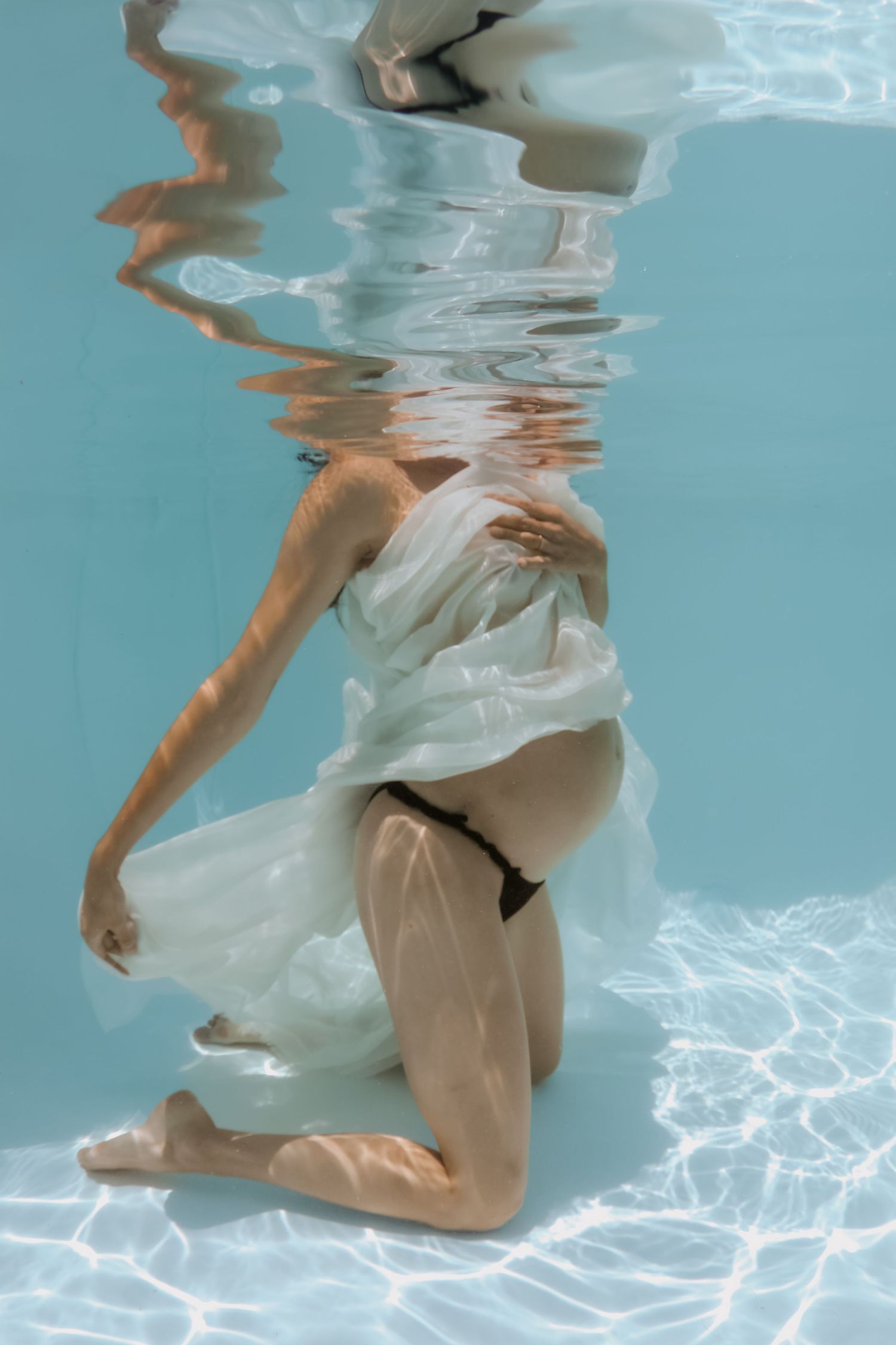 Pregnant woman kneeling underwater in a pool, draped in flowing white fabric, showcasing a maternity underwater photography session.
