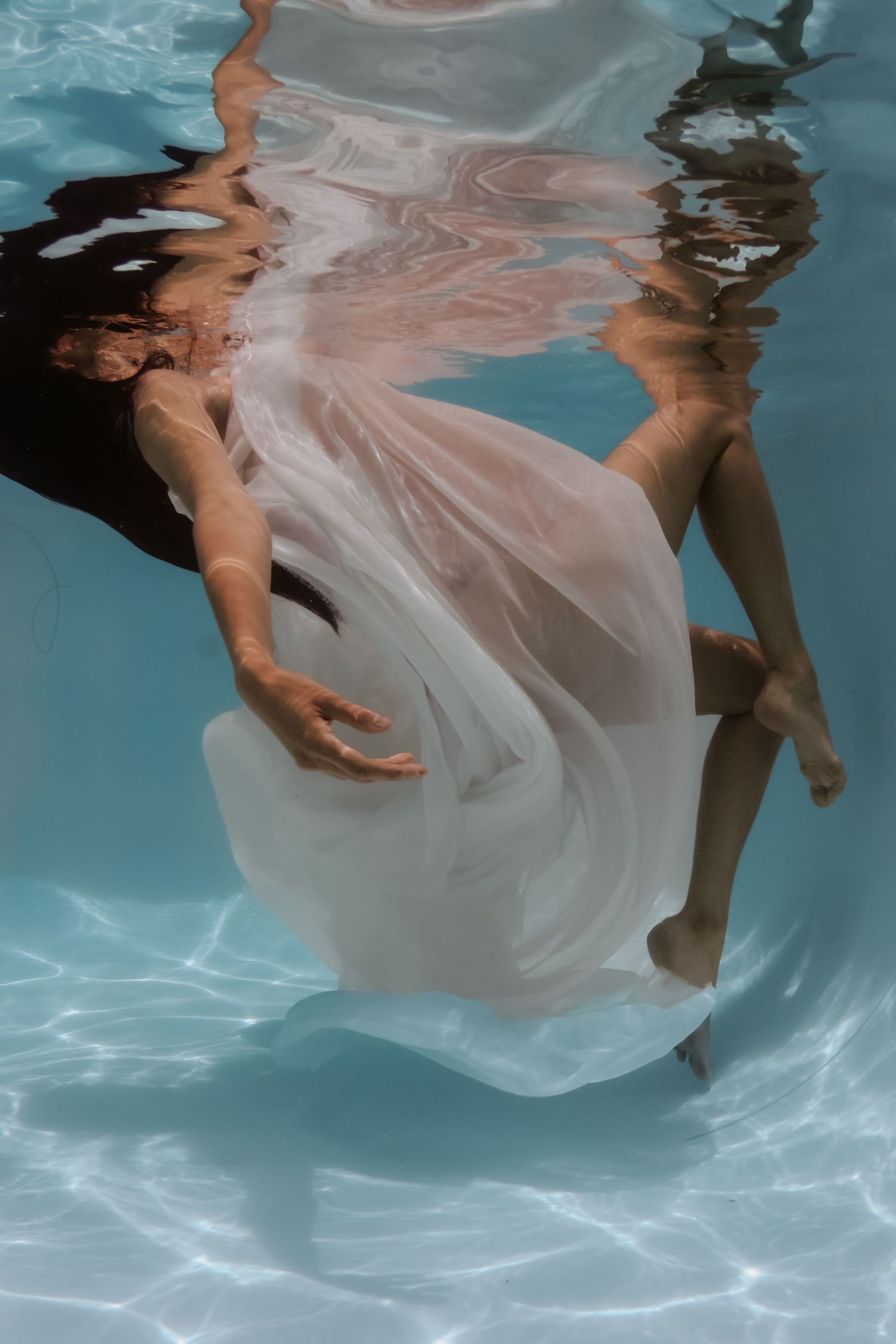 Ethereal underwater photo of a person in a flowing white dress submerged in a clear blue pool.