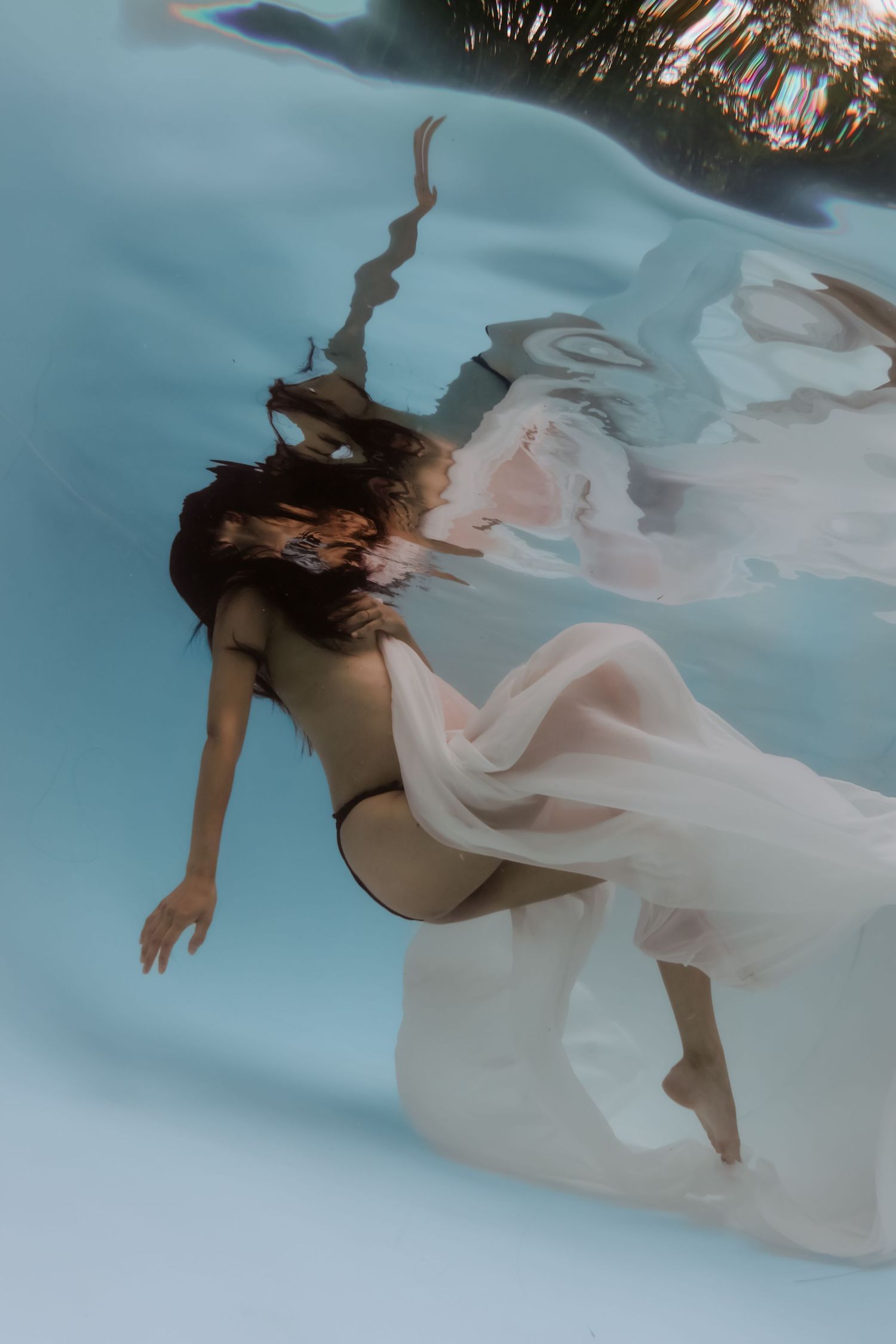 Pregnant woman in white flowing dress and black bikini floating gracefully underwater in a clear blue pool.