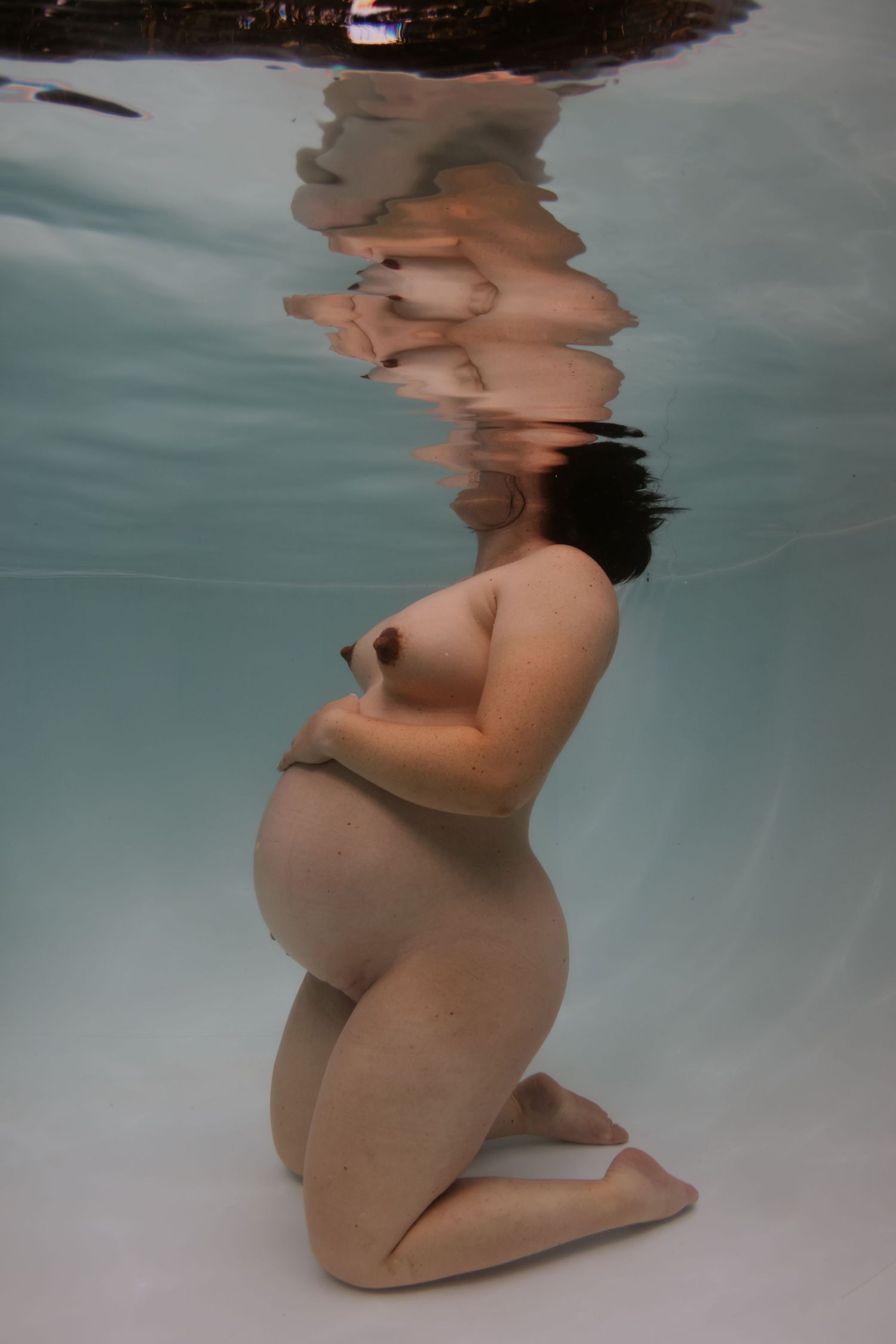 Pregnant woman kneeling underwater in a pool, captured in a serene underwater maternity photography session.