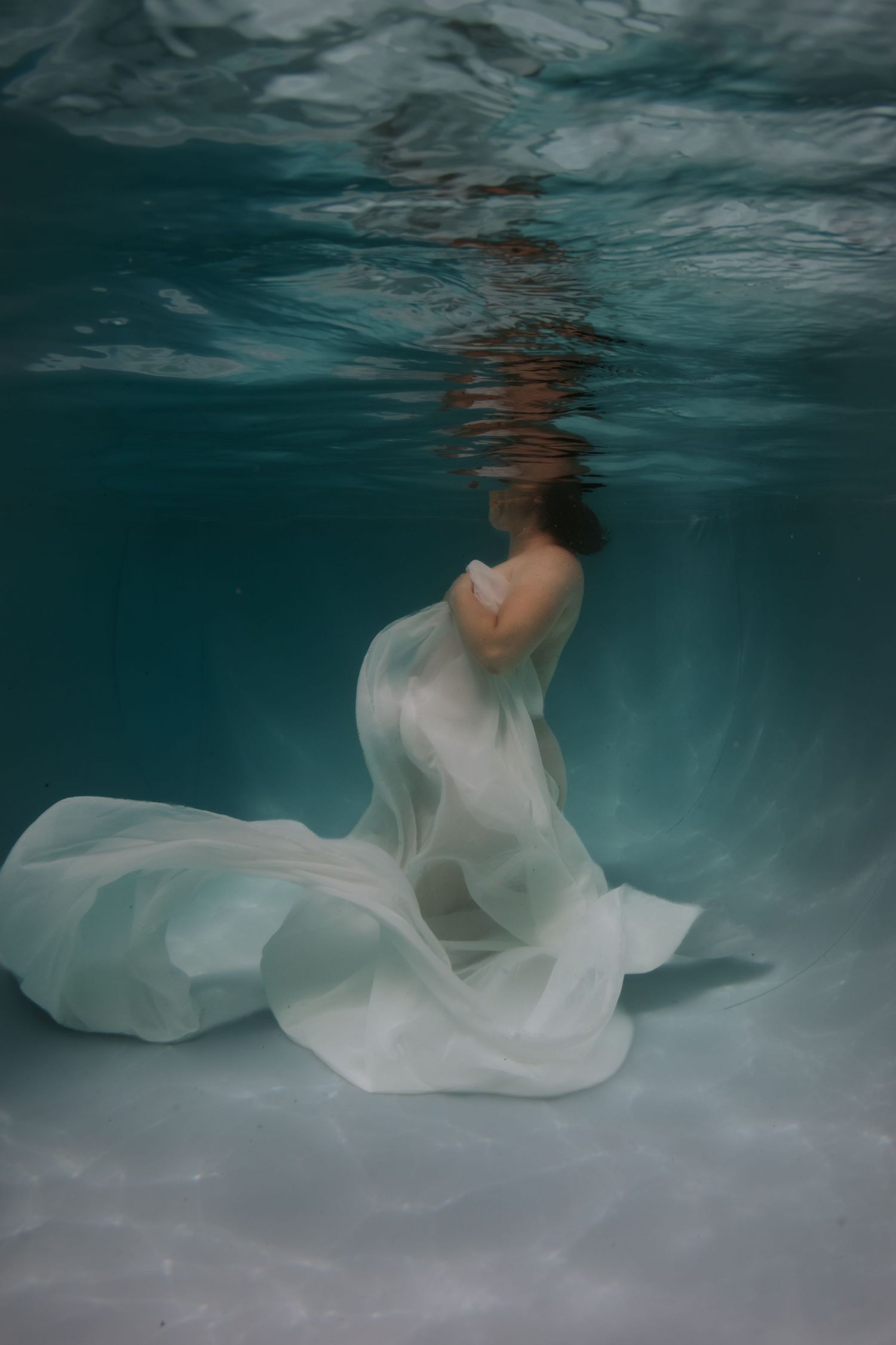 Ethereal underwater photo of a woman draped in flowing white fabric, submerged in clear turquoise water.