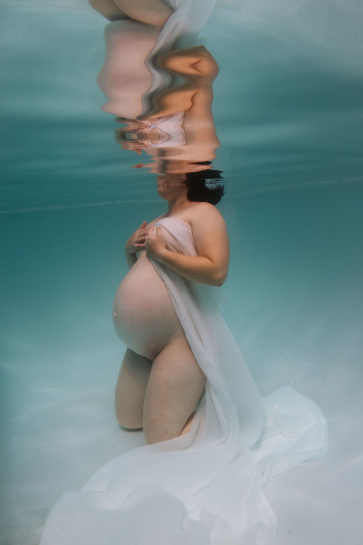 Pregnant woman kneeling underwater in white flowing fabric, serene maternity photography with aqua blue water.