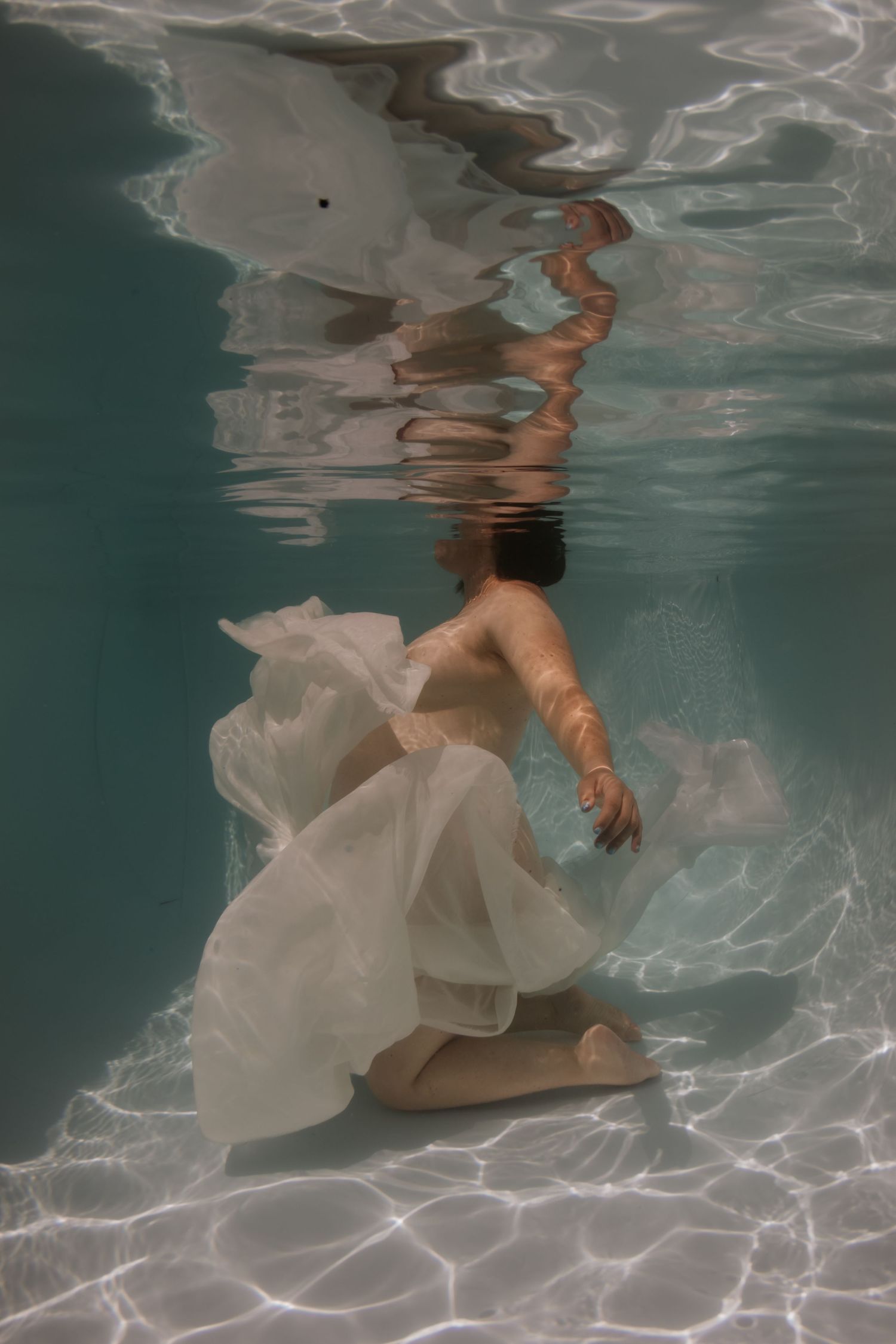 Person in white flowing dress submerged underwater in a pool, with light reflections on the surface above.
