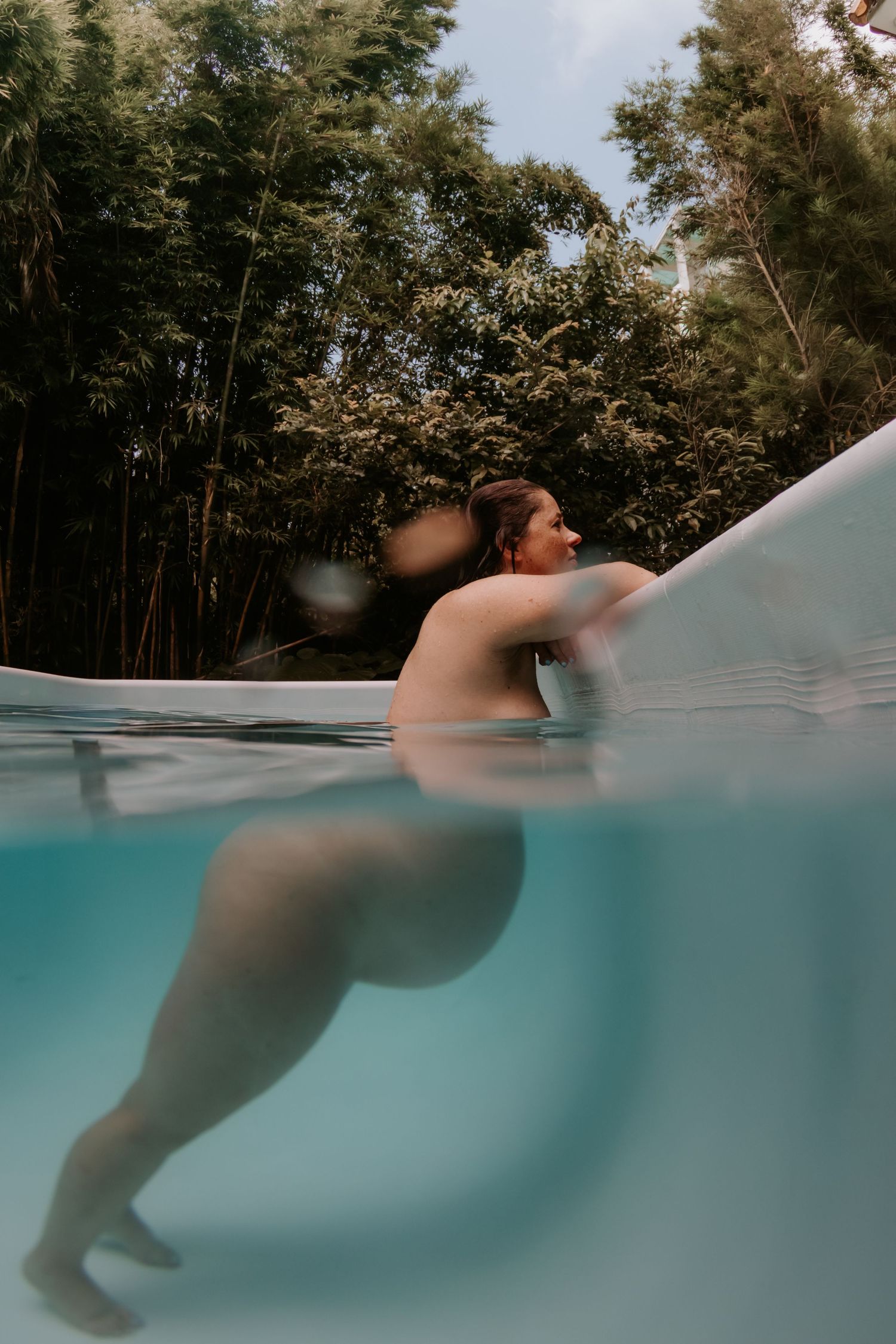 Pregnant woman relaxing in a clear pool surrounded by lush bamboo trees, captured in a split underwater view.