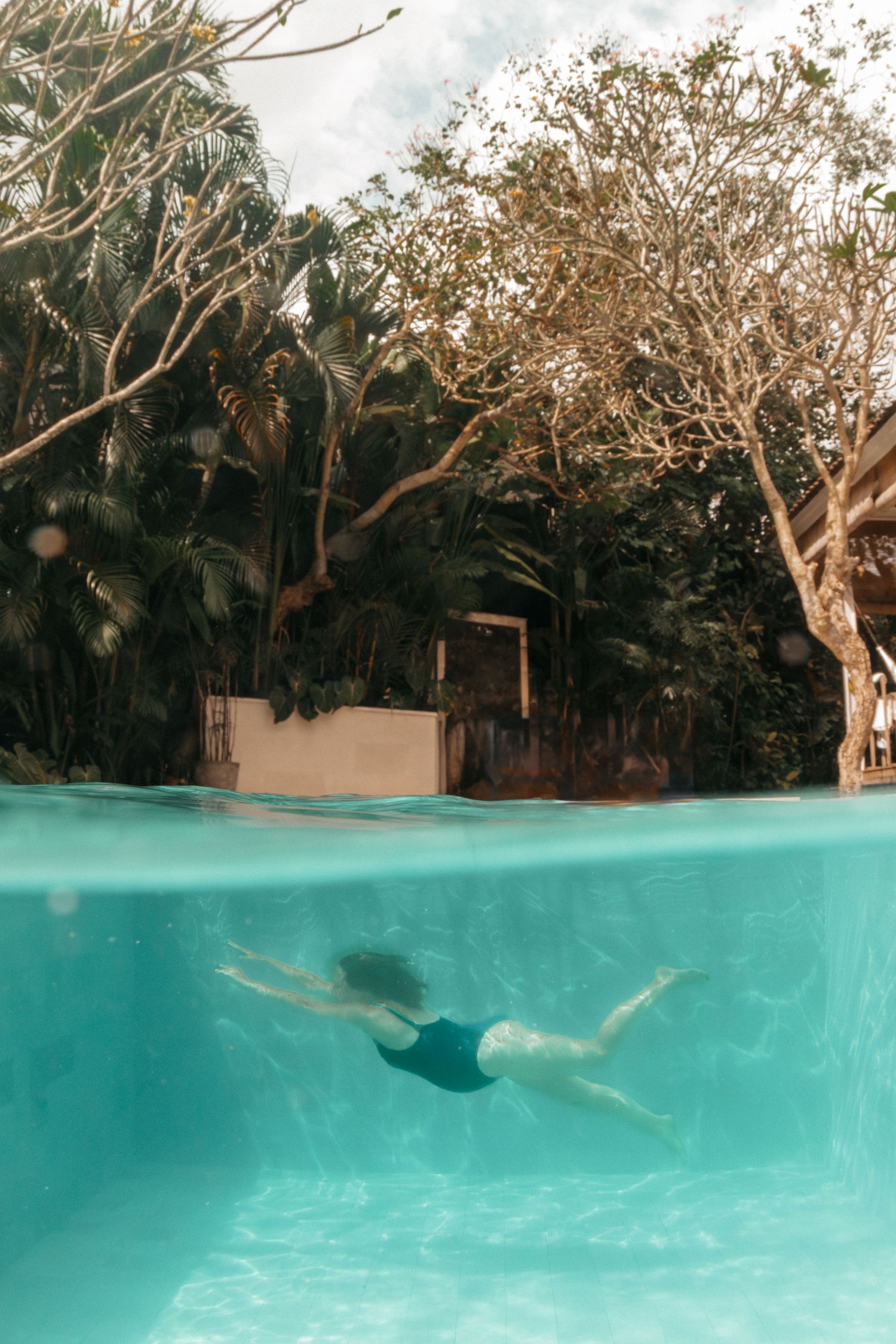 Split-view photo of a turquoise swimming pool showing someone swimming underwater with tropical trees visible above.