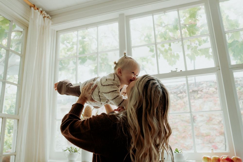 A mother joyfully lifts her smiling baby in the air near a bright window in a cozy home.