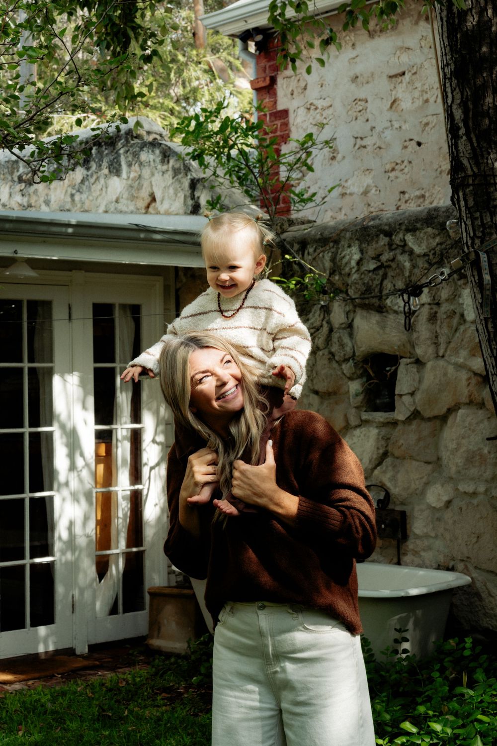 A smiling woman in a brown sweater carries a happy toddler on her shoulders outdoors near a rustic stone building.