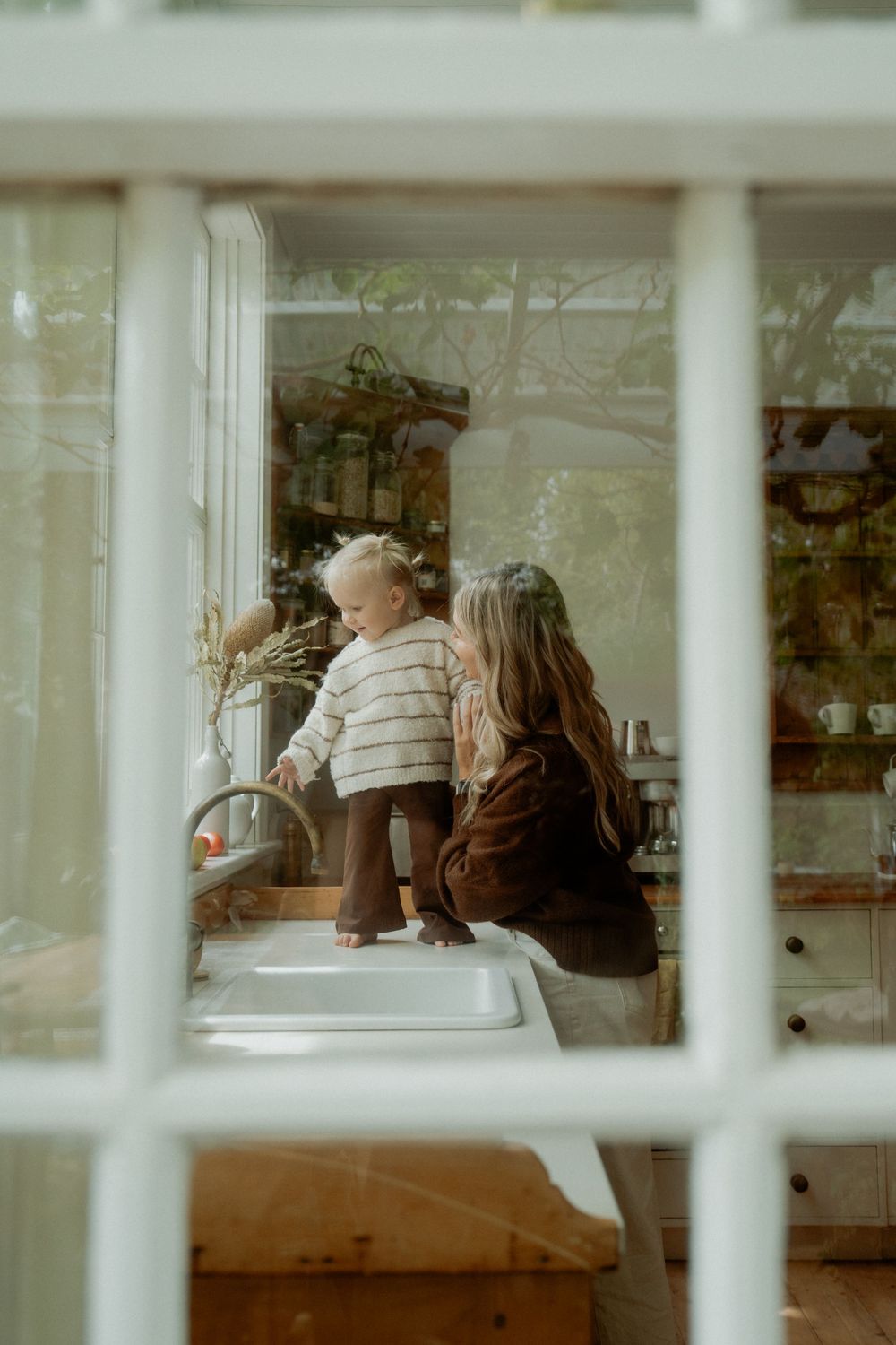 A toddler stands on a kitchen counter while a woman watches nearby, viewed through a window pane.