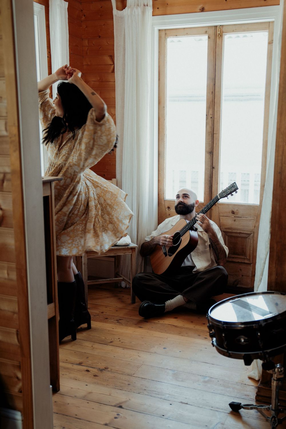 A woman dancing in a flowy dress while a bearded man plays acoustic guitar on the floor of a rustic wooden room.