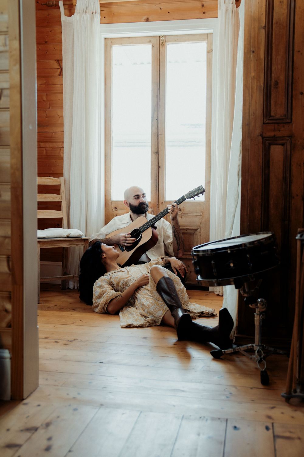 Bearded man playing acoustic guitar on wooden floor while woman listens in rustic log cabin room with drum kit nearby.