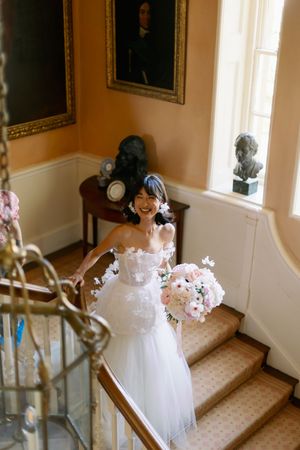 A joyful figure in a white tulle wedding dress stands on an elegant staircase with classic decor and floral arrangements.