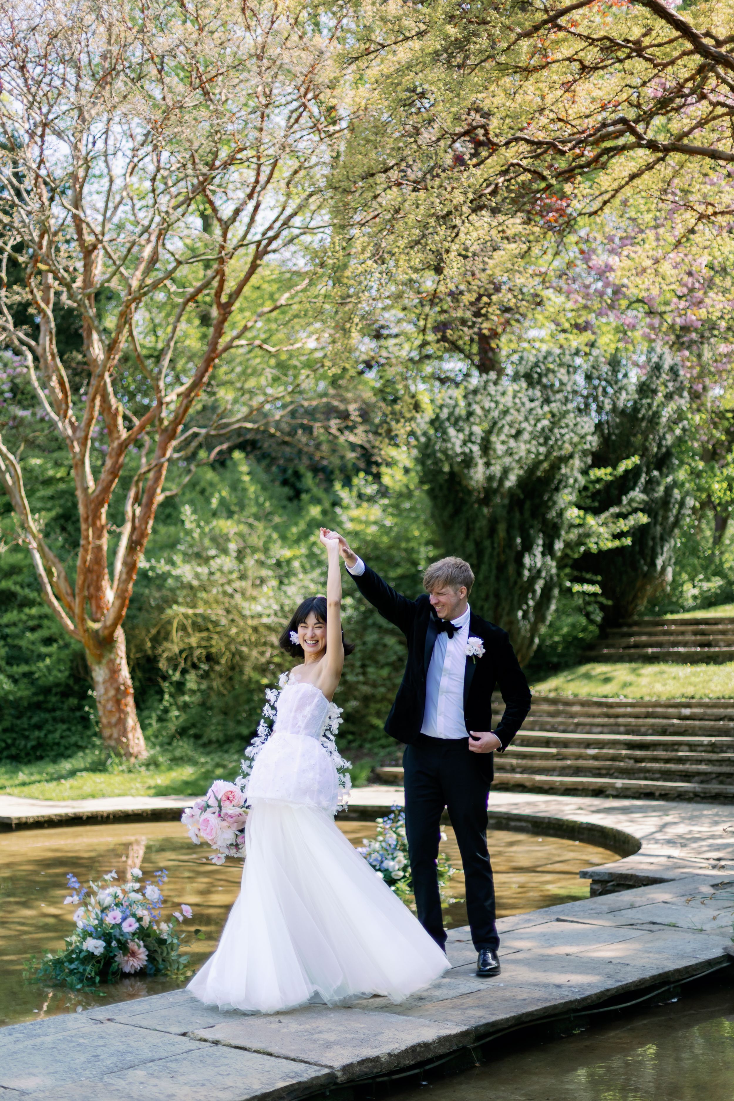 A couple in wedding attire pose joyfully by a garden pond surrounded by blooming trees and stone steps.