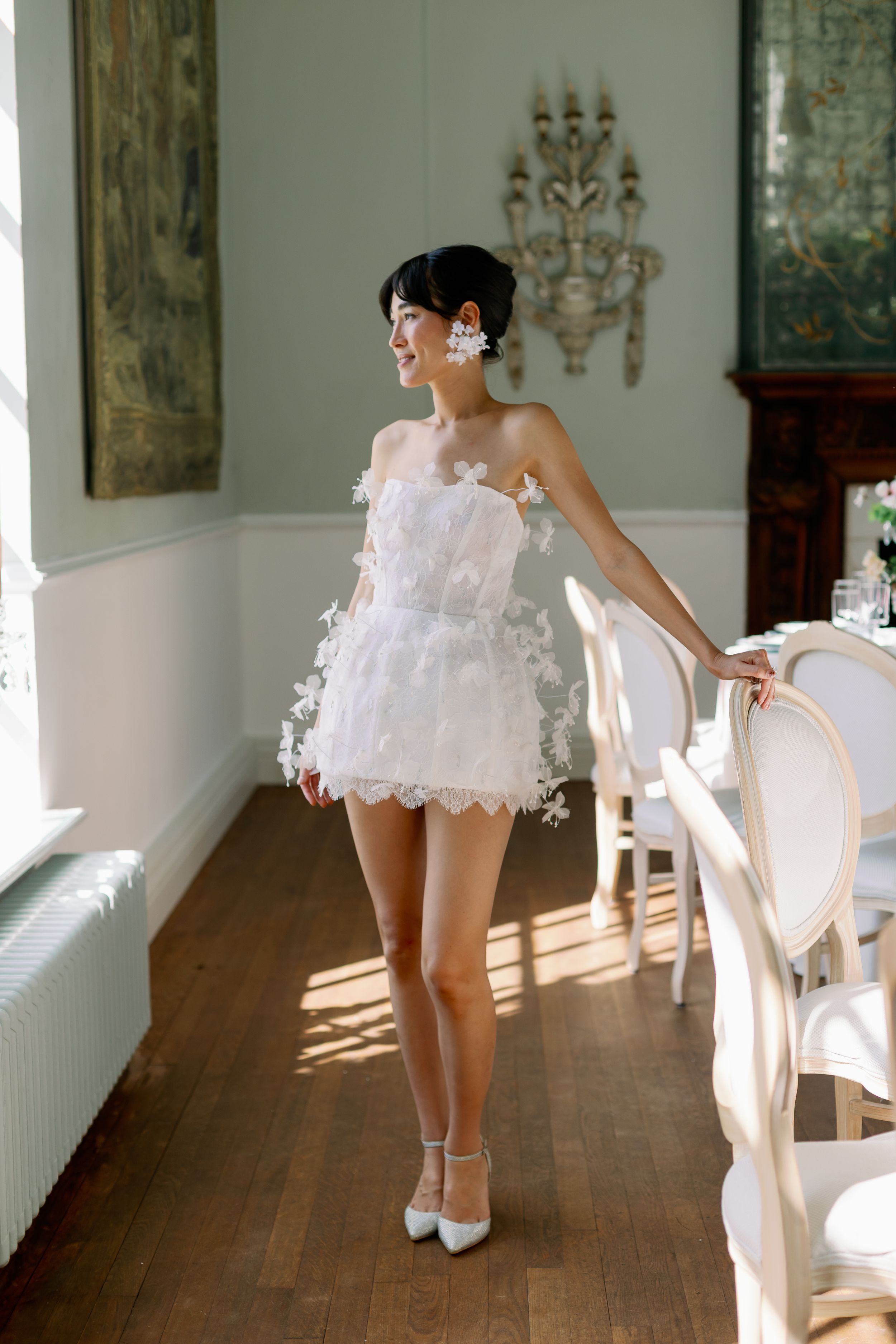 A white lace mini dress with floral appliques is styled with heels in an elegant room with hardwood floors and ornate decor.
